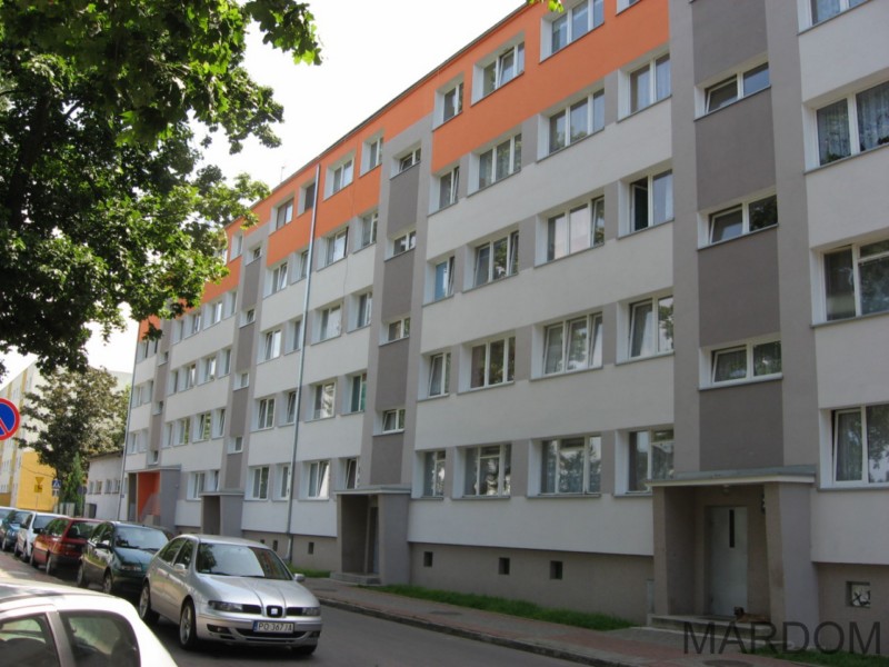 Odnowiony blok mieszkalny z ociepleniem, pomalowany w odcieniach szarości, bieli i pomarańczy, widoczny z poziomu ulicy z zaparkowanymi samochodami i zielenią drzew.